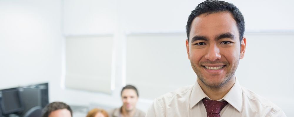 Docente sonriente en un aula de formación con otros asistentes al fondo.
